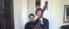 Dwóch mężczyzn uśmiecha się do zdjęcia. Pierwszy z nich, młodszy trzyma przed sobą kontrabas. Mężczyzna za nim jest ubrany na czarno i ma brodę.