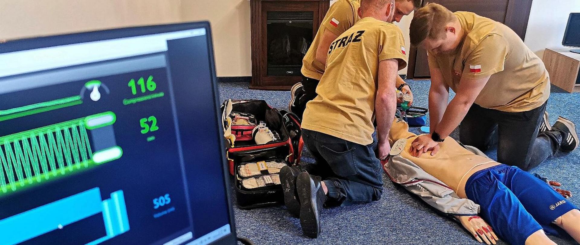 Zdjęcie zrobione w sali szkoleniowej podczas kursu kwalifikowanej pierwszej pomocy. Trzech strażaków ćwiczy udzielanie pierwszej pomocy na fantomie podłączonym defibrylatora. 