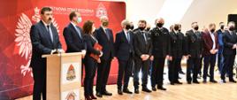 Mężczyzna stoi przy mównicy i przemawia obok niego inne osoby w garniturach dalej komendant wojewódzki w mundurze i inne osoby na tle loga PSP