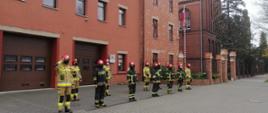 Pochmurno. Na tle budynku w kolorze ceglanym z dwiema brązowymi bramami stoją strażacy na baczność w ciemnych i piaskowych mundurach oraz czerwonych hełmach. Na twarzy mają czarne maseczki. Z prawej strony maszt z biało-czerwoną flagą. 