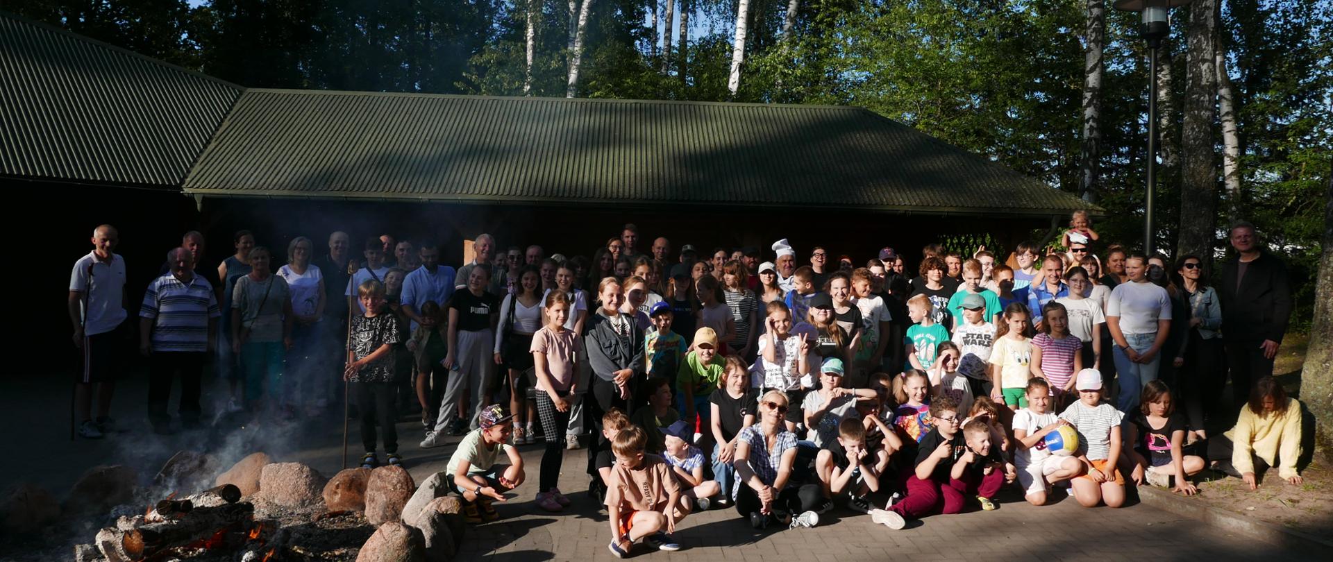 Las i specjalne miejsce do organizowania imprez. Na zdjęciu uczestnicy ogniska, dzieci, młodzież szkolna, nauczyciele i rodzice. Wszyscy uśmiechają się, obok palące się ognisko.