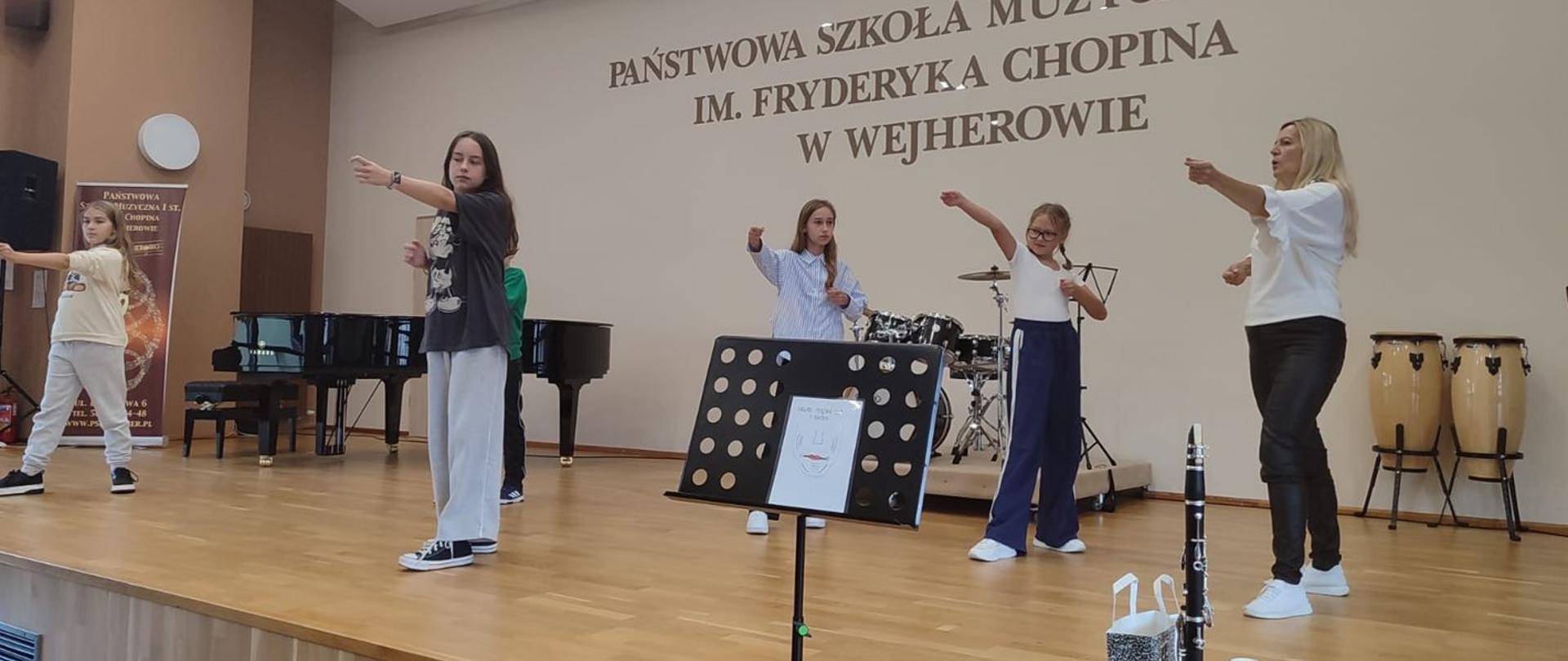 Na sali koncertowej pani Anna Łysko prowadzi warsztaty. Grupa uczennic wykonuje ćwiczenia zaprezentowane przez nauczycielkę. 