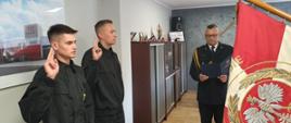Zbiórka strażaków w jasnym pomieszczeniu. Po prawej stronie widoczny biało - czerwony sztandar komendy. Po lewej dwaj funkcjonariusze w mundurach koszarowych składa ślubowanie. W środku komendant w mundurze wyjściowym odczytuje rotę ślubowania z granatowej teczki. W tle wyposażenie gabinetu.