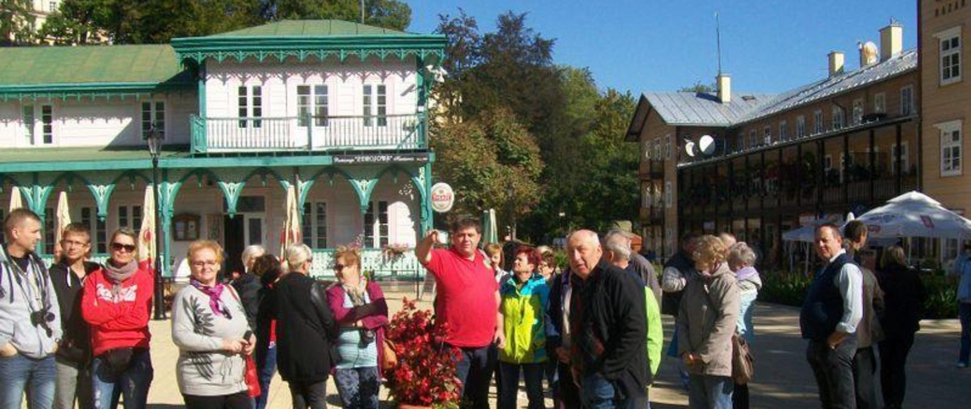 Na zdjęciu widzimy kobiety i mężczyzn zwiedzających stary rynek jakiegoś miasta. Osoby znajdują się w grupie, wyraźnie czymś zainteresowane, stoją na placu który jest utwardzony kostką brukową koloru piaskowego . W tle widzimy j zabytkowe budynki kamienic koloru białego i brązowego, zielone dachy.