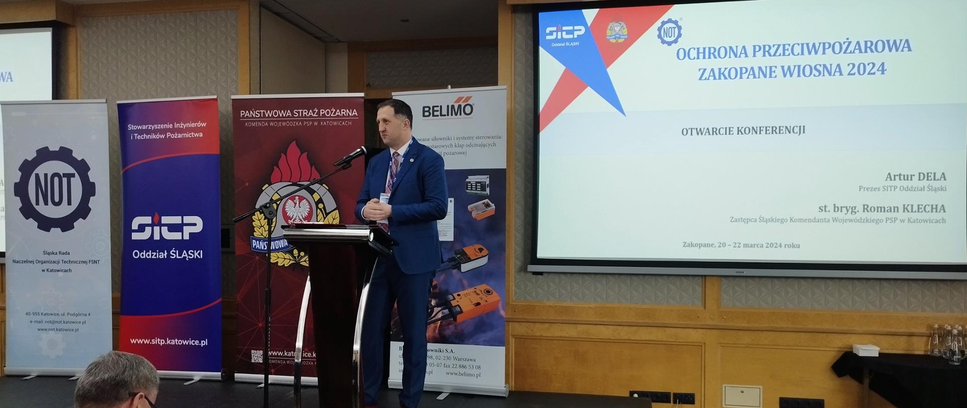 Na zdjęciu mężczyzna ubrany w garnitur stoi przed mikrofonem na podwyższeniu przed publicznością, za nim rozciągnięte roll-upy, z tyłu na rzutniku widoczny napis z nazwą konferencji 