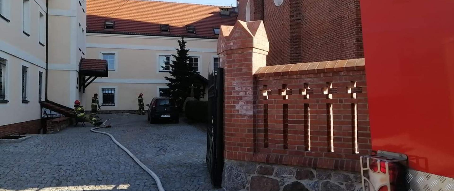 Zdjęcie przedstawia dziedziniec klasztoru Franciszkanów w Radziejowie oraz strażaków z rozciągniętą linią gaśniczą, podczas gaszenia pożaru.
