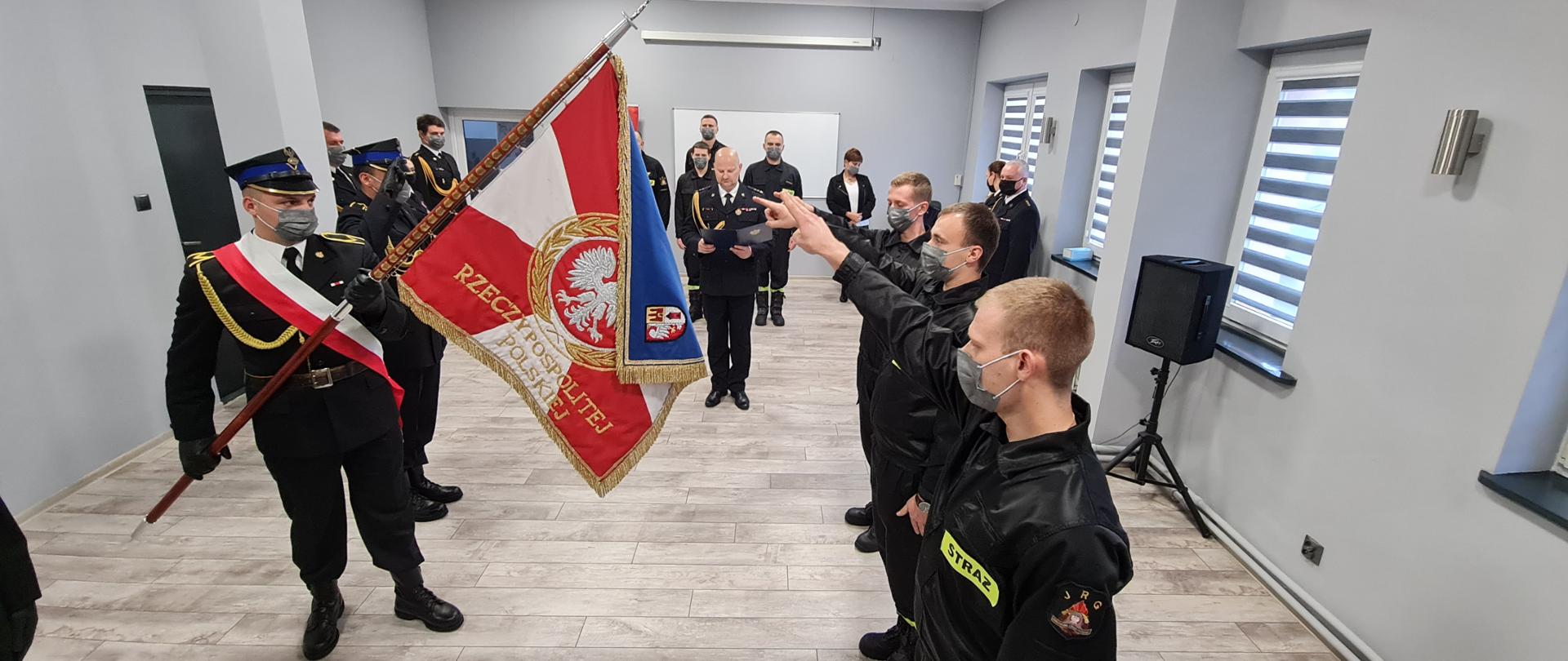 Trzech nowoprzyjętych strażaków stoi z wyciągniętymi prawymi rękami w kierunku sztandaru KP PSP w Żyrardowie. W tle stoi komendant powiatowy wraz z pracownikami komendy.