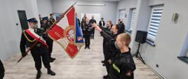 Trzech nowoprzyjętych strażaków stoi z wyciągniętymi prawymi rękami w kierunku sztandaru KP PSP w Żyrardowie. W tle stoi komendant powiatowy wraz z pracownikami komendy.