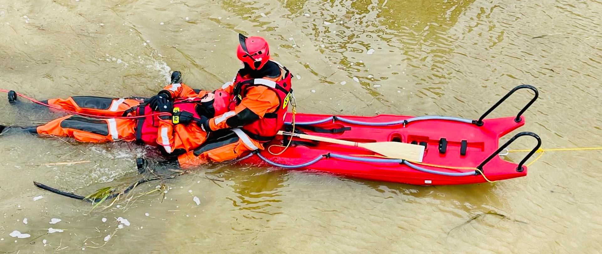 Zdjęcie pokazuje strażaka na saniach lodowych, który transportuje do brzegu poszkodowanego. Strażacy ubrani w specjalistyczny ekwipunek na tle wody.