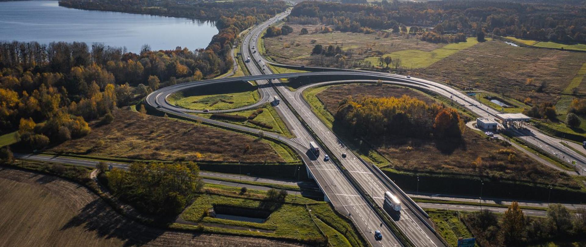 Widok z lotu ptaka na węzeł drogowy z estakadą i pętlami zjazdów nad wielopasmową trasą; po drogach jadą samochody, wokół rozciągają się pola, łąki i lasy w jesiennych barwach, a w tle widoczne jest jezioro.