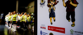 Dzieci ubrane w kamizelki odblaskowe stoją i śpiewają, obok stoi baner z logiem kampania dziewczynka i chłopiec wraz z logami instytucji zaangażowanych w projekt edukacyjny.