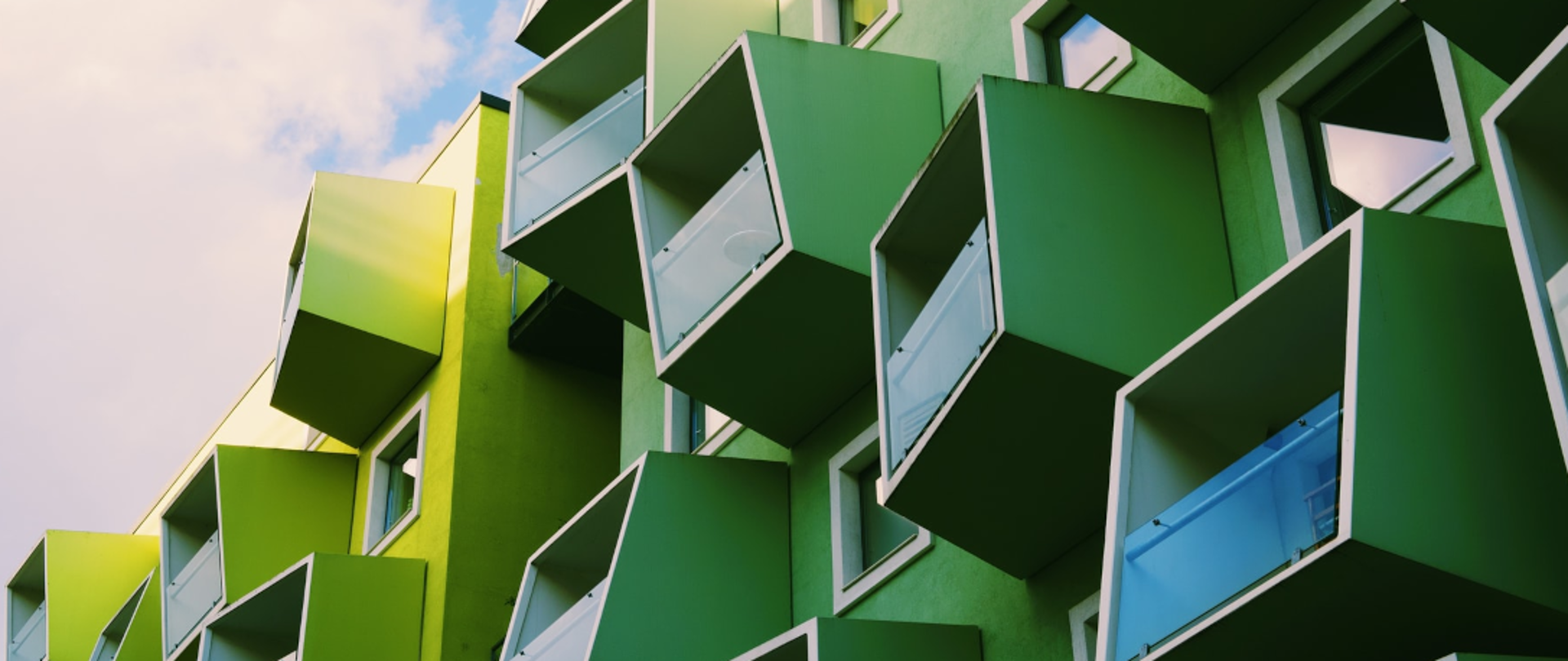 Zielony budynek z nowoczesnymi, sześciennymi balkonami i szklanymi balustradami na tle pochmurnego nieba