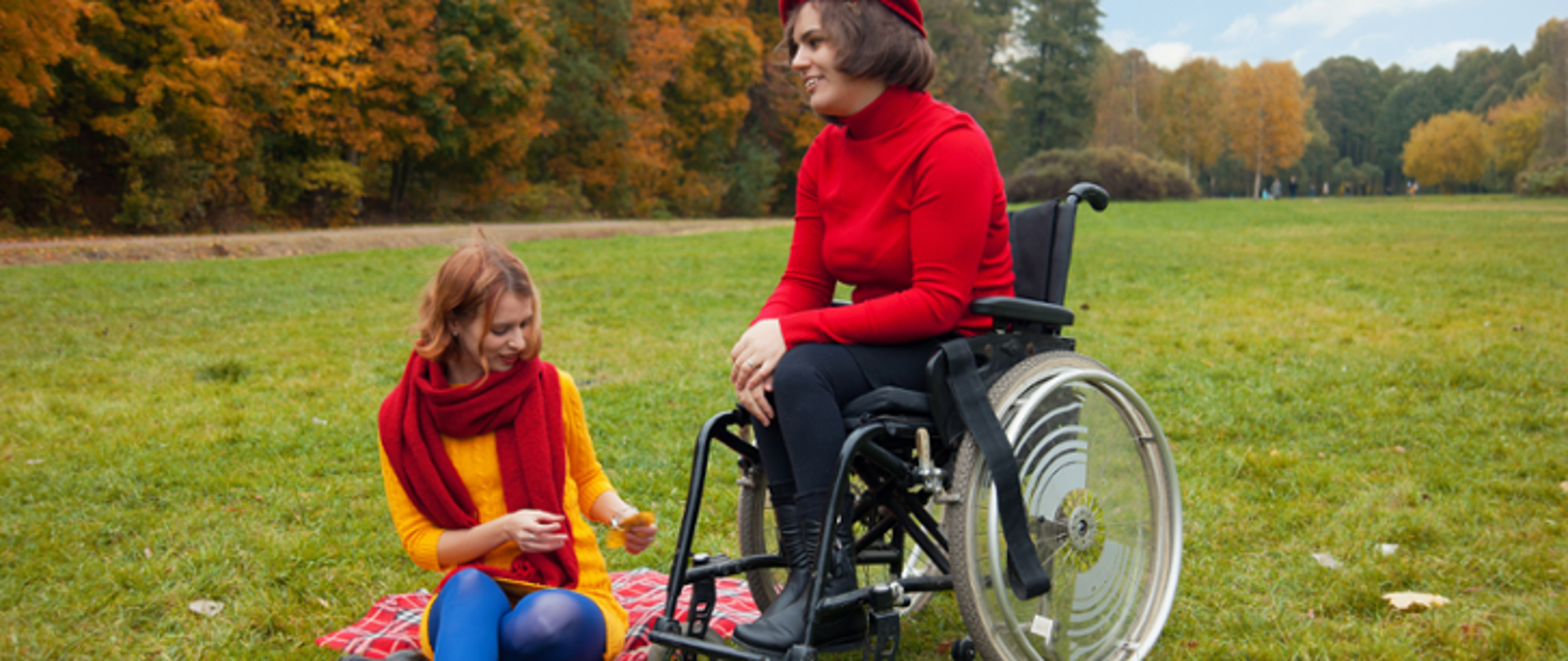 Kobieta z widoczną niepełnosprawnością ruchową na polanie w parku. Obok na kocu siedzi druga kobieta. 