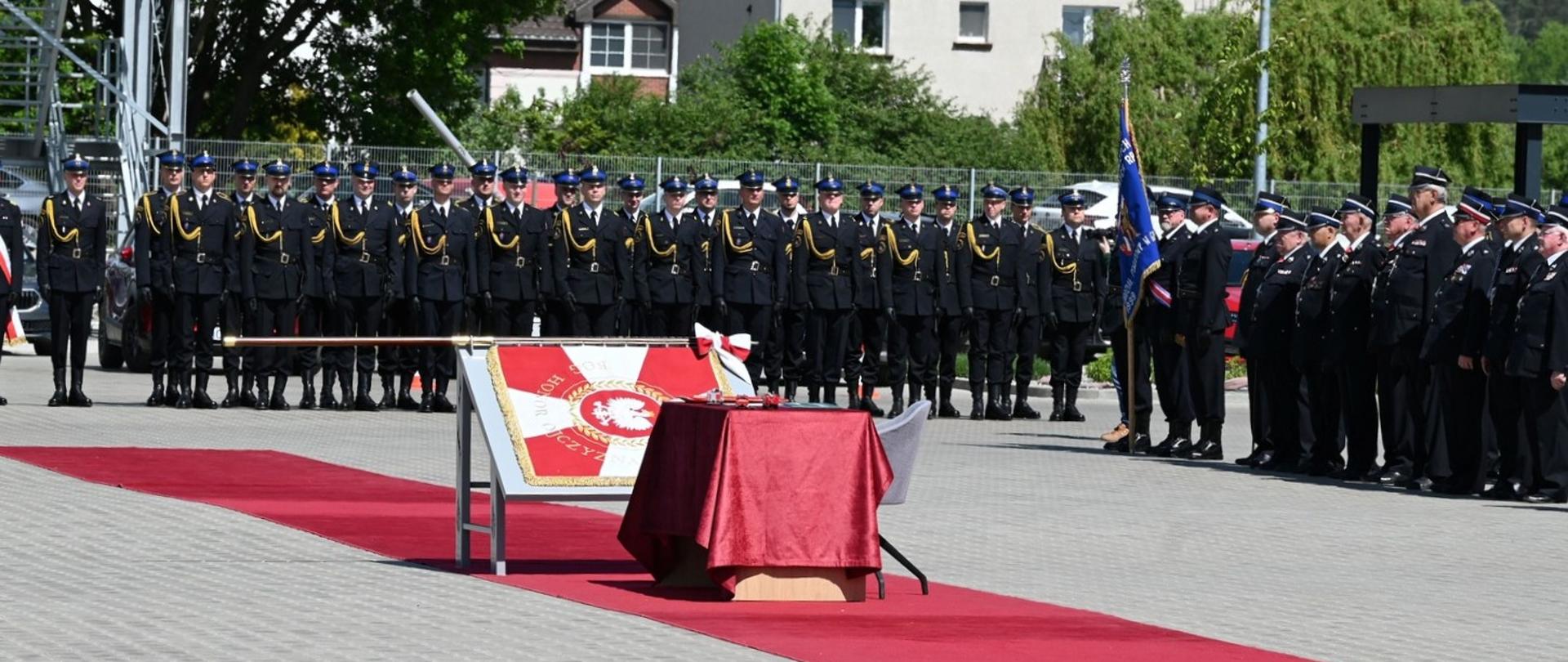 Uroczysty apel strażacki na placu przed budynkiem, z rzędami umundurowanych strażaków, czerwonym dywanem i sztandarem z godłem Polski.