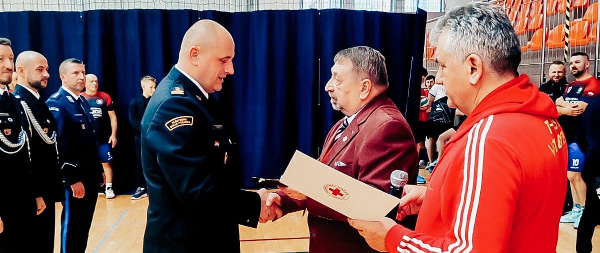 Zdjęcie przedstawia komendanta powiatowego PSP w Ustrzykach Dolnych odbierającego nagrody od związku PCK. Widać również innych zgromadzonych na turnieju.