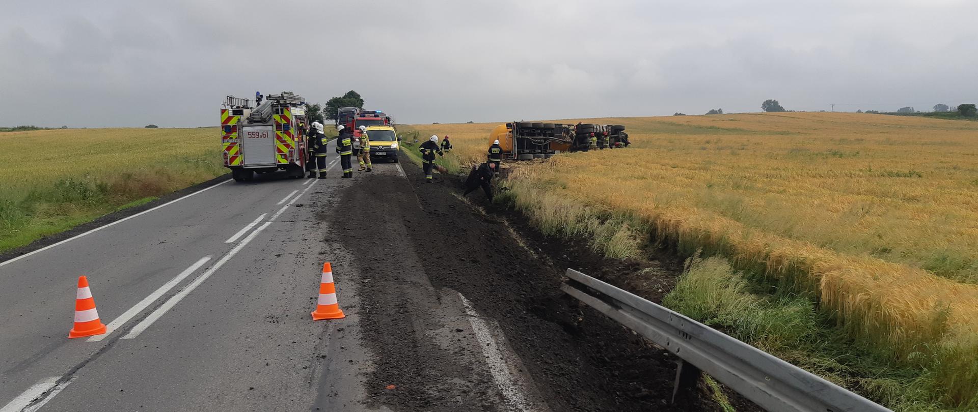 Zdjęcie przedstawia samochód ciężarowy leżący na lewym boku z cysterną do przewozu cementu, na drodze krajowej widoczne samochody bojowe i pachołki odgradzające miejsce zdarzenia