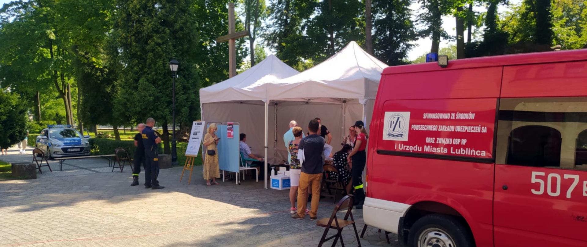 Na zdjęciu widoczna jest aleja w parku. Rozłożone dwa namioty z mobilnymi punktami szczepień przeciwko COVID. Przy namiotach stoją ludzie, druh OSP i policjant. Po prawej stronie stoi czerwony samochód OSP.