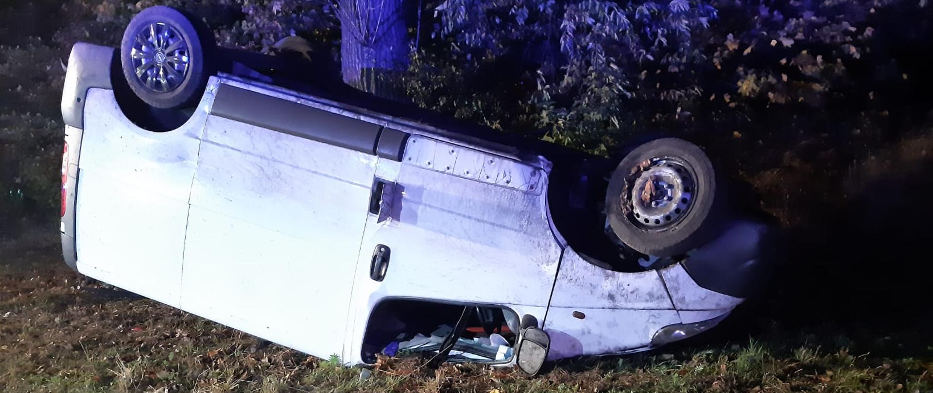 Na zdjęciu znajduje się biały samochód typu bus. Samochód znajduje się poza drogą wojewódzką na poboczu. Samochód znajduje się na dachu, prawdopodobnie kierujący stracił panowanie nad pojazdem i uderzył w drzewo a następnie dachował.