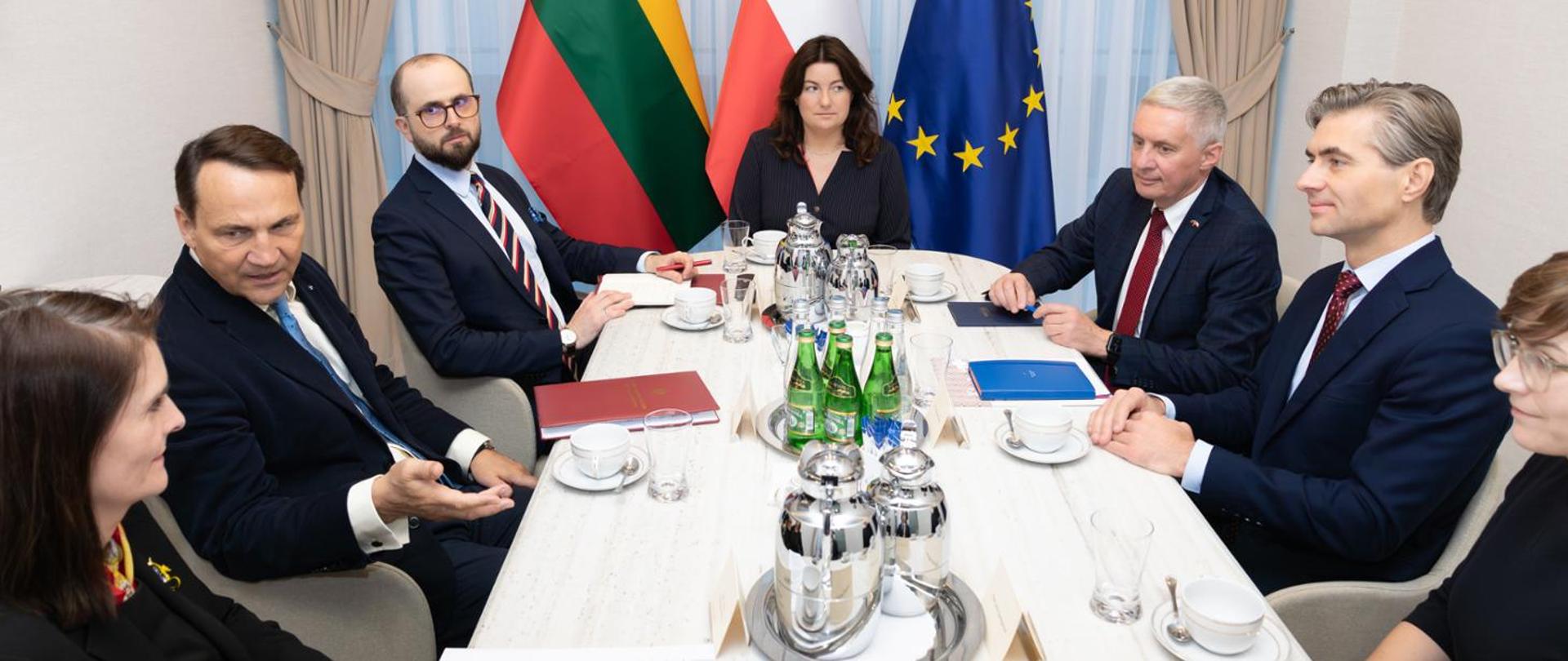 Na zdjęciu jest siedem osób - czterech mężczyzn w garniturach i trzy kobiety ubrane w ciemne stroje. Wszyscy siedzą przy owalnym stole, na którym znajdują się butelki z wodą, szklanki i notatniki. W tle widać flagi: Litwy, Polski i Unii Europejskiej. 