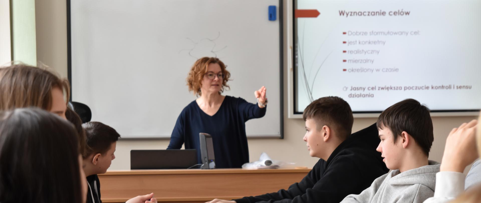 Nauczycielka prowadzi wykład w klasie szkolnej, stojąc przed ekranem z prezentacją dotyczącą motywacji, podczas gdy uczniowie siedzą w ławkach i uważnie słuchają.