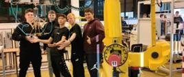 Grupa pięciu młodych mężczyzn stoi przy dużym, żółtym ramieniu robota przemysłowego w hali technologicznej.