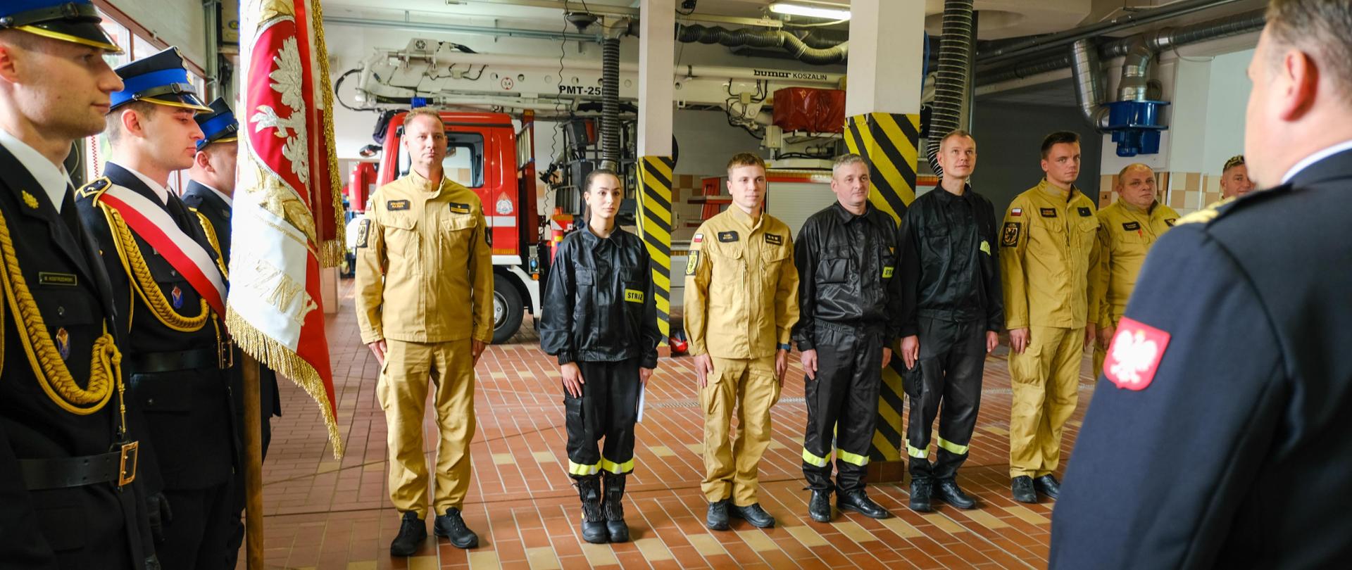 Strażacy w umundurowaniu galowym i bojowym stoją w szeregu podczas uroczystości w remizie. Na pierwszym planie widoczny sztandar z godłem Polski