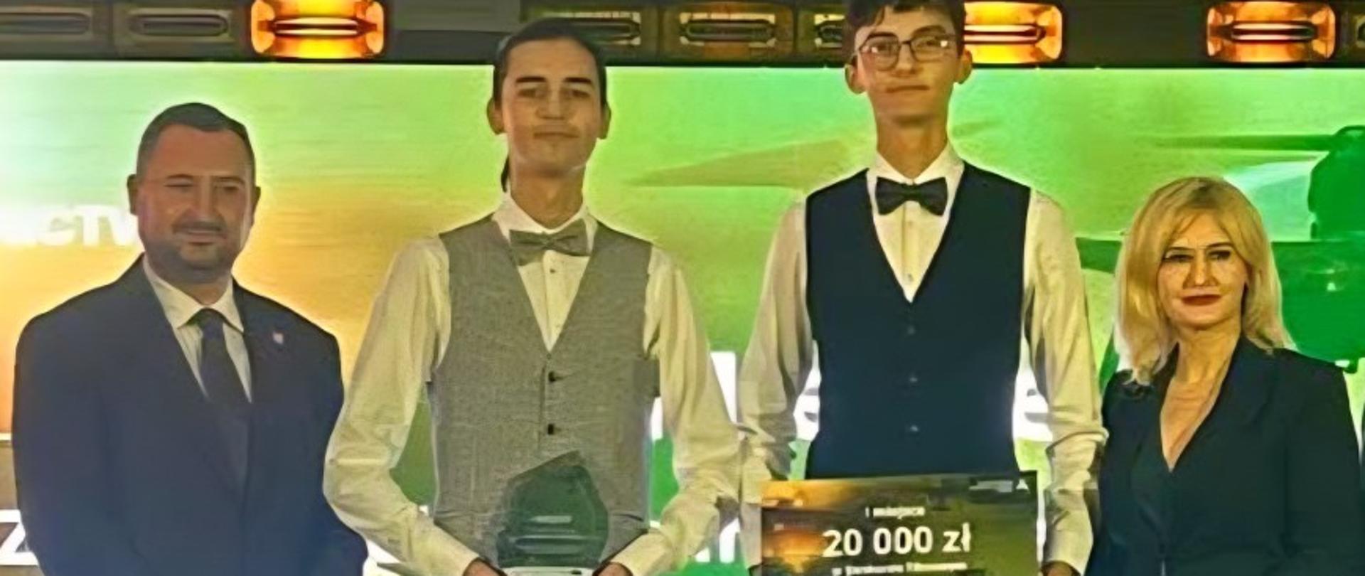 Cztery osoby stoją na scenie podczas wręczenia nagród, jedna z nich trzyma statuetkę, a druga czek