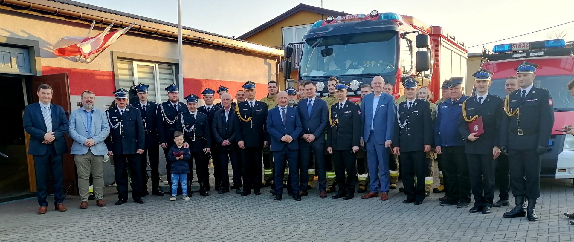na zdjęciu grupa osób i strażaków za nimi dwa samochody strażackie oraz budynki