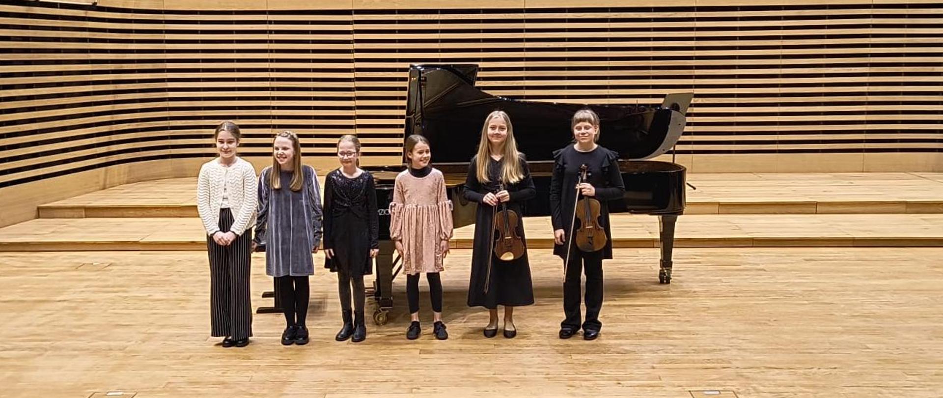 Sześć dziewczynek stoi na scenie, dwie trzymają skrzypce.