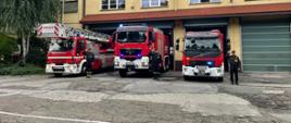 Trzy samochody pożarnicze stojące przed bramami garażowymi JRG. Widoczne włączone niebieskie sygnały świetlne. Przy każdym z samochodów stoi strażak w umundurowaniu koszarowym. Strażacy salutują.