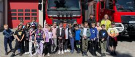 Grupa dzieci szkolnych stoi przed dwoma czerwonymi wozami strażackimi na tle budynku straży pożarnej. Dzieci uśmiechają się i pozują do zdjęcia wraz z dwiema osobami dorosłymi. W tle widoczne są czerwone drzwi garażowe i okna budynku. Pogoda jest słoneczna.