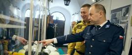 Na zdjęciu podczas wernisażu wystawy „Na STRAŻY bezpieczeństwa mieszkańców Ziemi Gorlickiej" 2 osoby ogląda eksponaty strażak w ubraniu służbowym koloru pisakowego i komendant powiatowy policji w ubraniu galowym koloru granatowego. W tle widoczne zdjęcia i eksponaty pożarnicze pompa ręczna, działko, hełmy, aparat powietrzny.