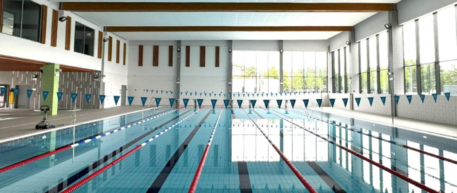 Kryta pływalnia z kilkoma torami, nad którymi wiszą trójkątne flagi; po prawej stronie duże okna, po lewej zielony filar i wejście.

