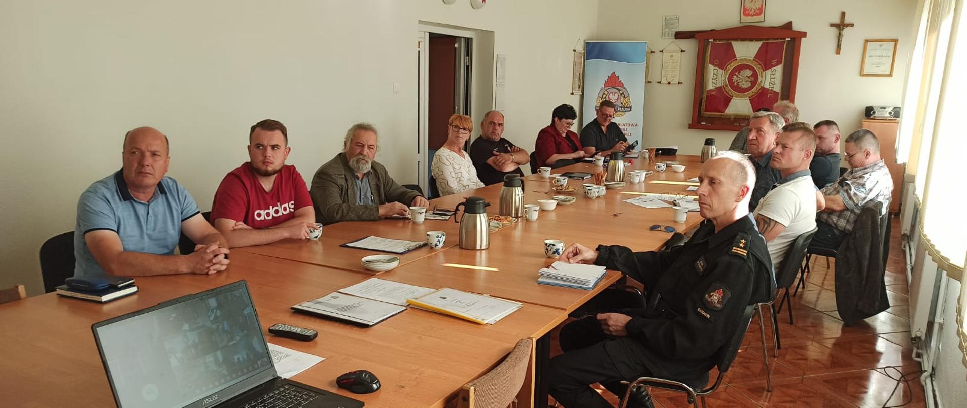 Zdjęcie przedstawia sale wykładową na której odbywa się wideokonferencja. Przy stole siedzą osoby biorące udział w wideokonferencji a na planszy wyświetlany są inne grupy.