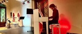 Zdjęcie przedstawia ucznia grającego na pianinie podczas koncertu. W tle czerwone światła. Pianino koloru białego.