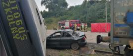 Autobus przewrócony na samochód osobowy na przejeździe kolejowym, niedaleko lokomotywowa oraz strażacy. 