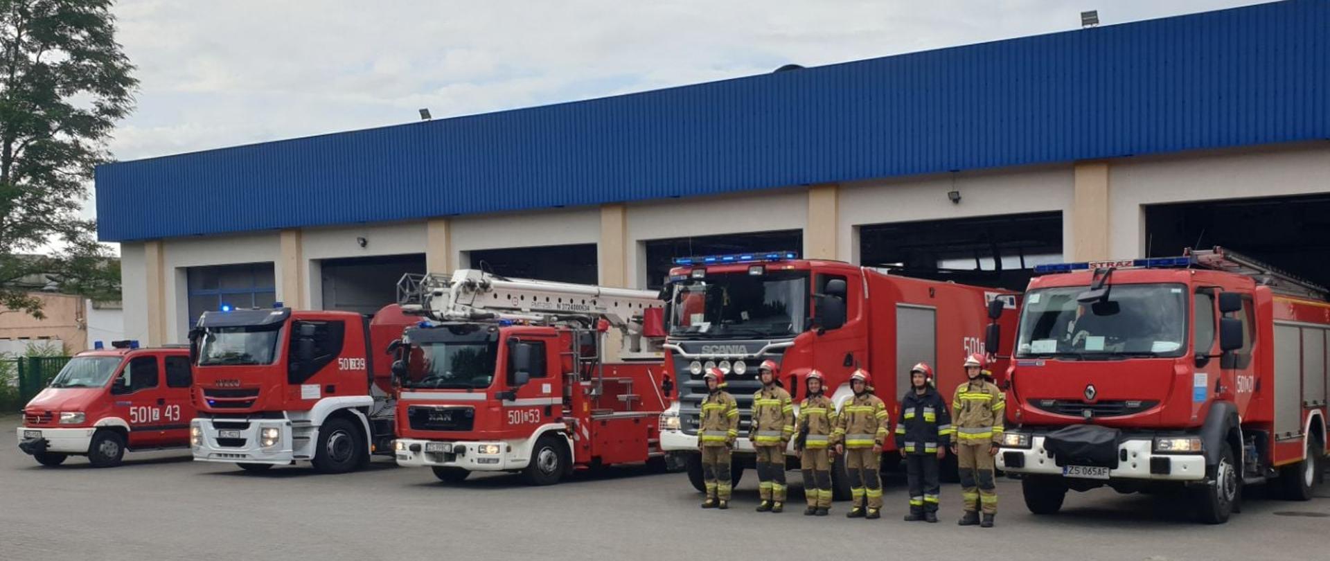 Na zdjęciu widoczny plac z polbruku z napisem Straż i fragment budynku strażnicy. Przed garażami budynku stoi pięć strażackich pojazdów z właczonymi sygnałami świetlnymi. Przed nimi stojący w równym szeregu, wyprostowani strażacy, w piaskowych ubraniach bojowych i w czerwonych hełmach na głowie. Drugi od prawej w czarnym ubraniu bojowym.