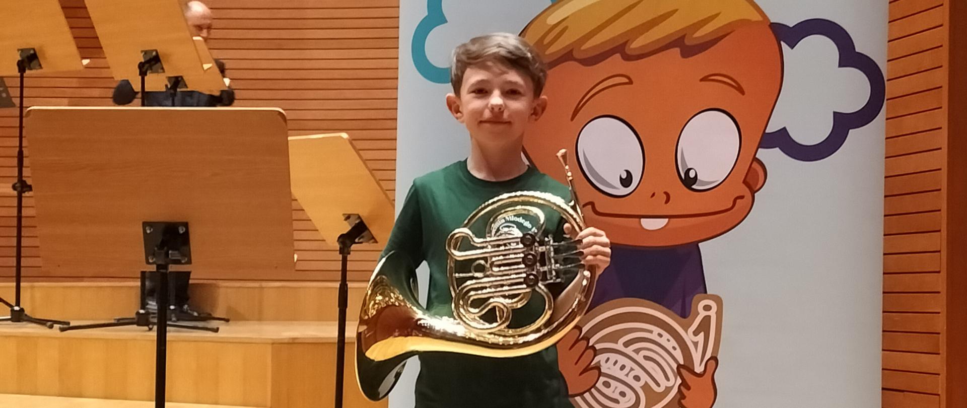 Kolorowe zdjęcie Tomasza Tompalskiego stojącego na scenie i trzymającego w rękach waltornie, za nim znajduje się kolorowy baner reklamujący Akademię Młodego Waltornisty