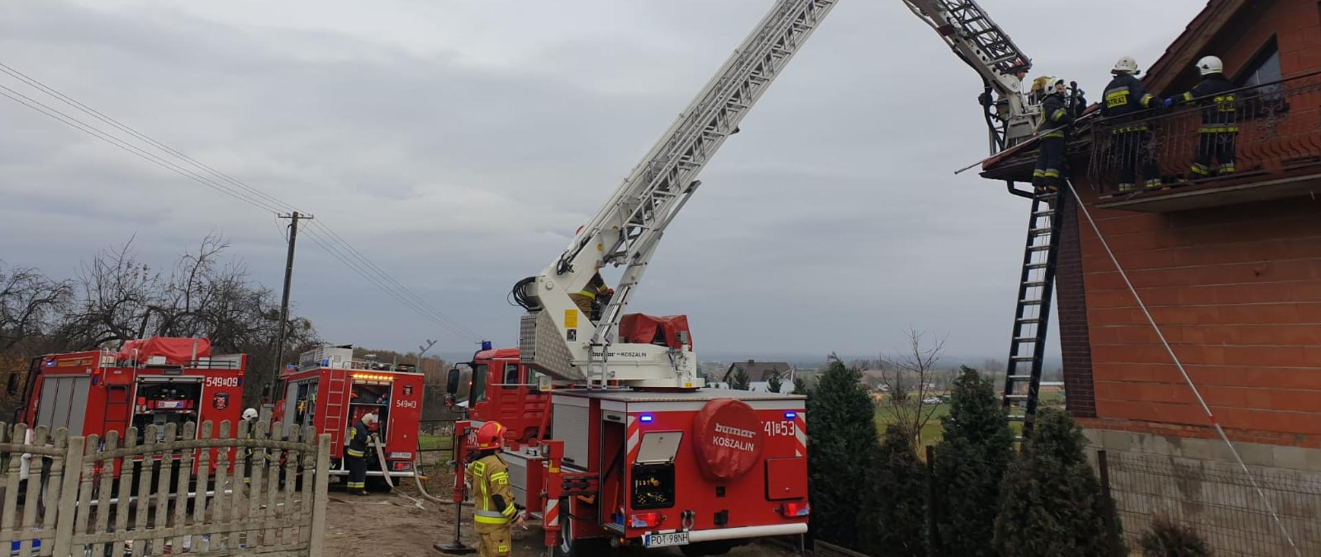 Zdjęcie przedstawia działania ratowniczo - gaśnicze podczas pożaru dachu budynku jednorodzinnego. Na zdjęciu widać strażaków działających na wysokości z kosza SH 23, którzy dokonują częściowej rozbiórki pokrycia dachowego w celu zlikwidowania zagrożenia a także 2 zastępy OSP podczas działań. Zdjęcie wykonane w porze dziennej przed posesją.