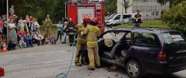 Zdjęcie przedstawia strażaków podczas pokazu z użyciem narzędzi hydraulicznych oraz wraku samochodu - strażacy odcinają dach samochodu. W tle widać samochód straży pożarnej oraz uczestników pikniku.
