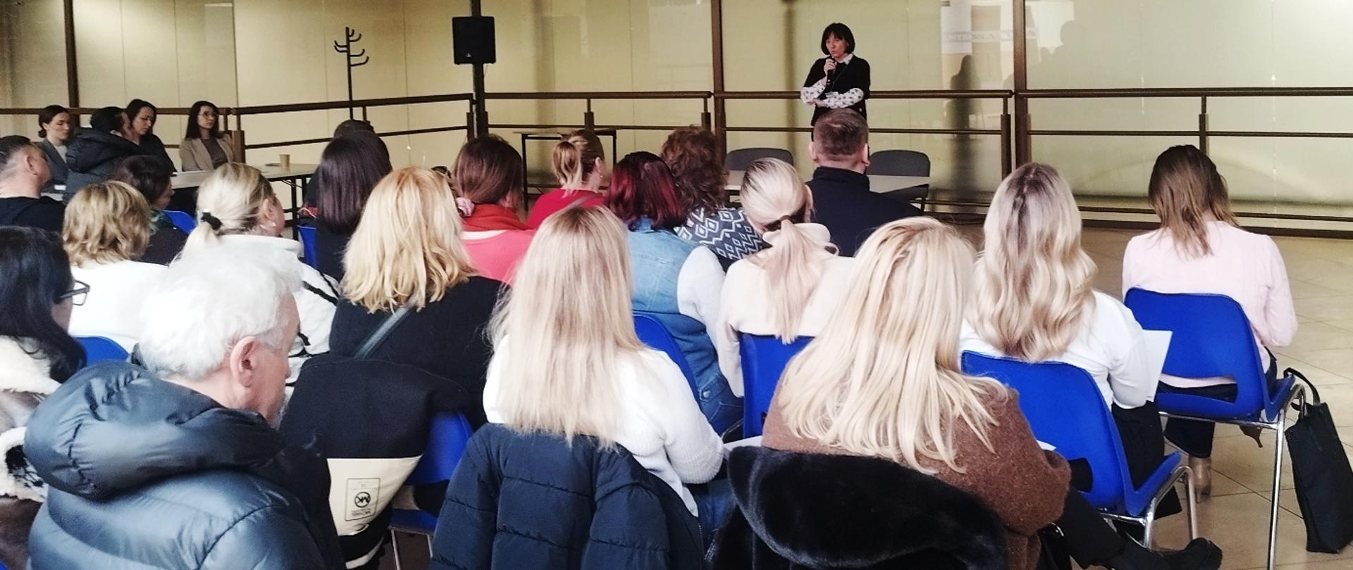 Grupa osób siedzi na niebieskich krzesłach i słucha kobiety przemawiającej na małej scenie w jasnej sali konferencyjnej