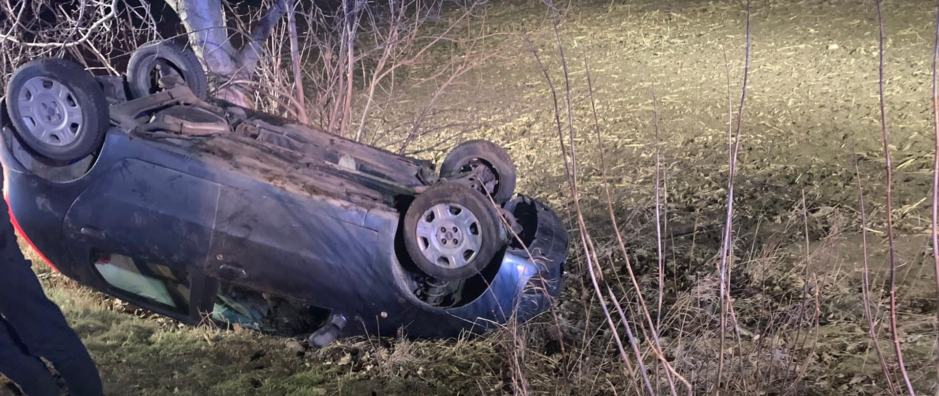 samochód osobowy leży na dach pomiędzy nim widoczne krzaki oraz drzewo. 