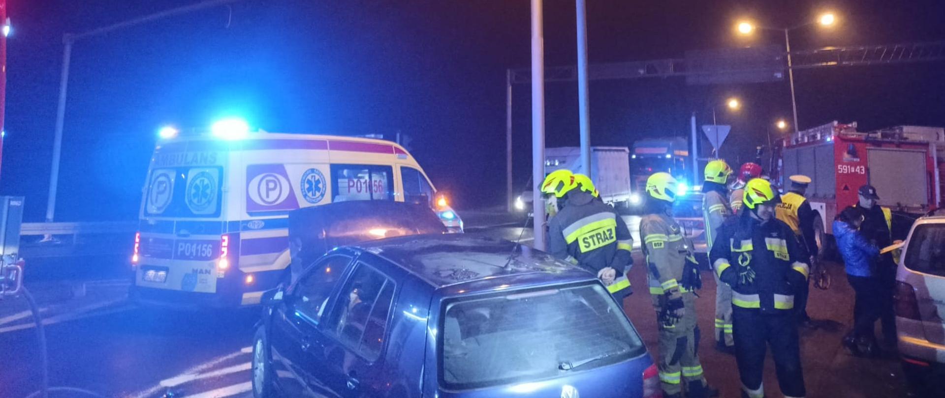 Widać uszkodzony samochód na skrzyżowaniu, za nim karetka pogotowia, po prawej strażacy, jest ciemno