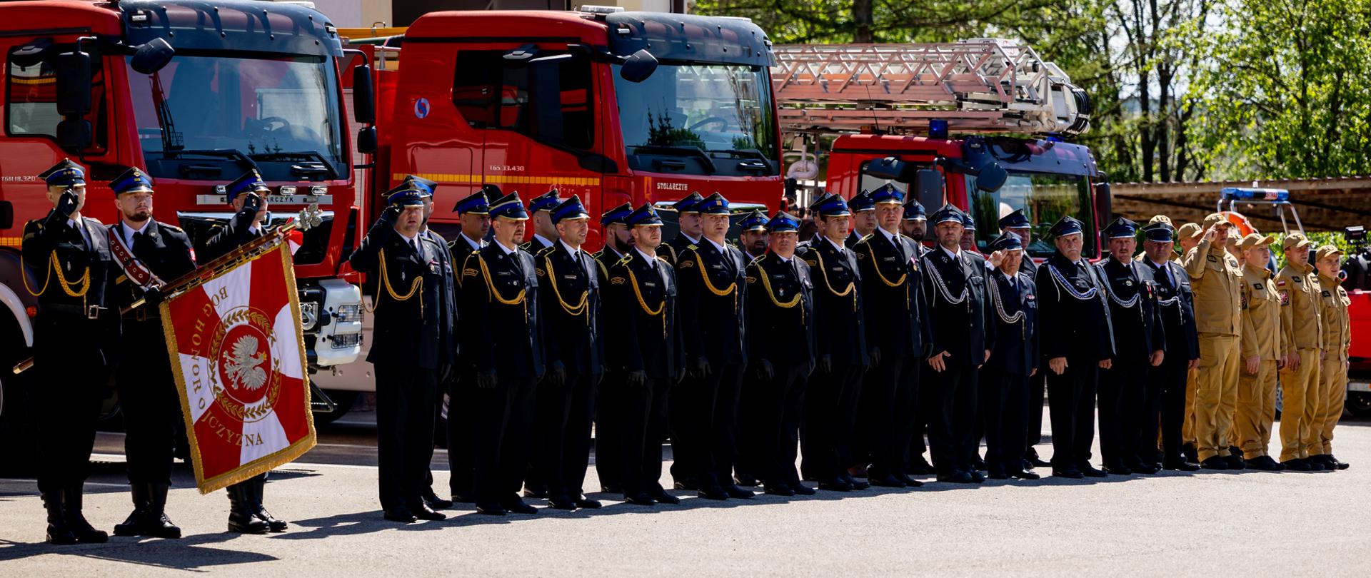 Pododdział strażaków i członków Młodzieżowej Drużyny Pożarniczej stoi w szyku podczas uroczystej zbiórki przed budynkiem komendy.