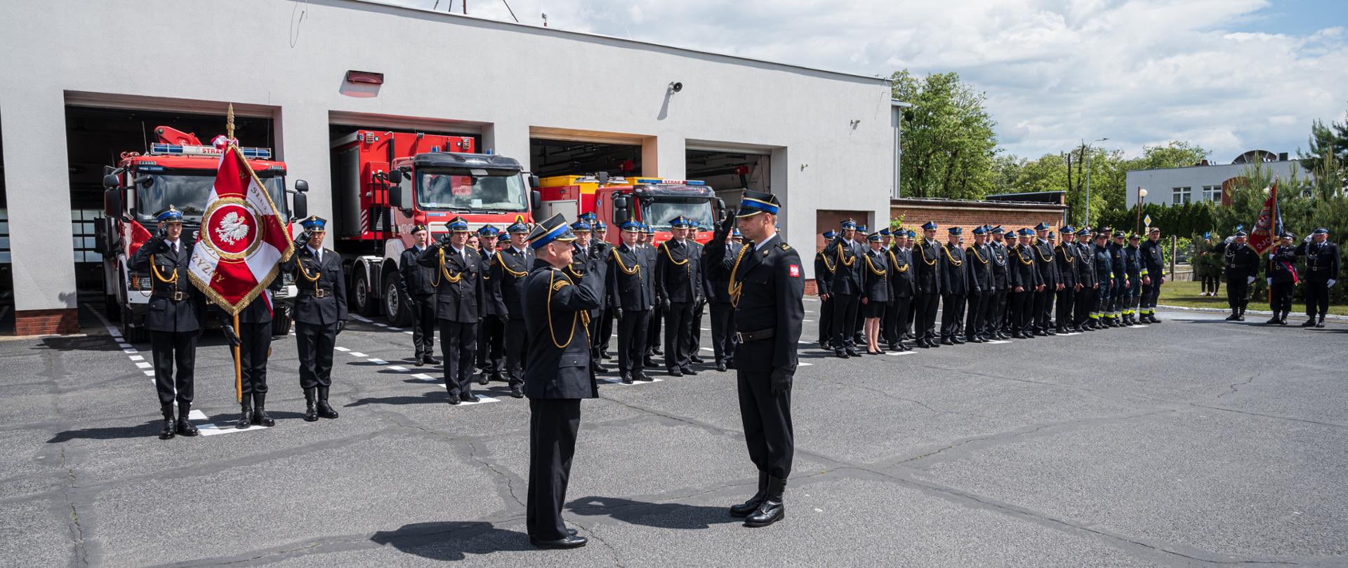 Dwóch strażaków salutuje do siebie w tle strażacy stoję w rzędzie