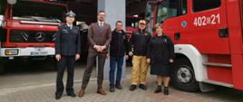 Strażacy Państwowej Straży Pożarnej przedstawiciele gminy oraz druhowie ochotniczej straży pożarnej stoją przed garażami obok samochodu gaśniczego.