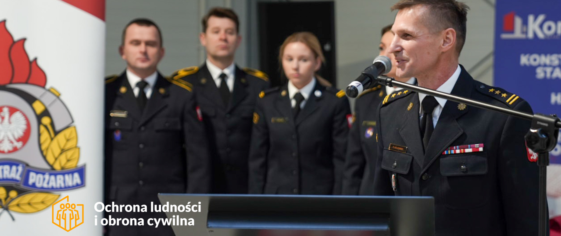 Strażak w mundurze wyjściowym stoi przy mikrofonie, za mównicą. W tele kilkoro strażaków, a z lewej strony fragment banera firmowego z logo PSP.