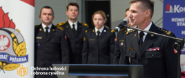 Strażak w mundurze wyjściowym stoi przy mikrofonie, za mównicą. W tele kilkoro strażaków, a z lewej strony fragment banera firmowego z logo PSP.