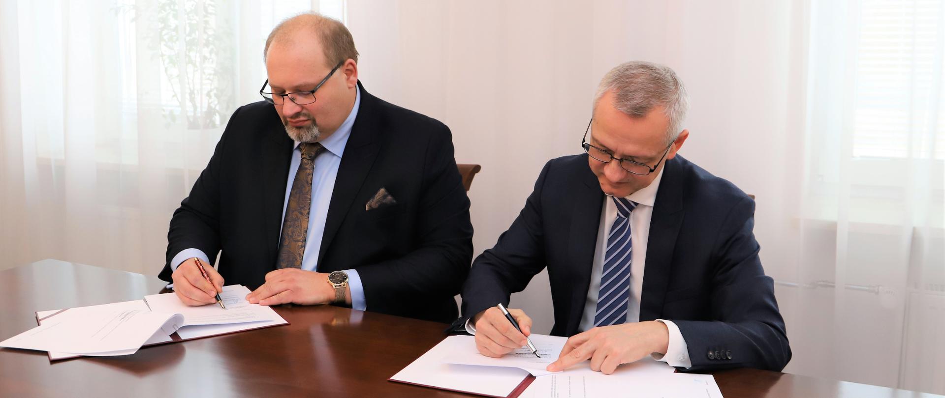 Andrzej Cieślak - prezes DYNACON Sp. z o.o. i Marek Zagórski - minister cyfryzacji siedzą przy stole i podpisują umowę o współpracy.