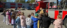 Zdjęcie przedstawia strażaka który stoi na placu jednostki przed podnośnikiem hydraulicznym i opowiada dzieciom o pracy strażaka